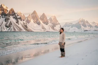 Femme debout sur une plage arctique des îles Lofoten en Norvège sous le soleil de minuit estival, avec des montagnes enneigées en arrière-plan